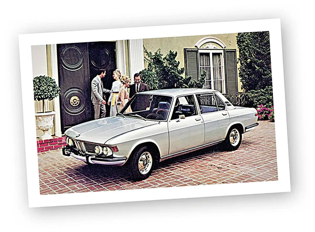 Elegancki, srebrny BMW E9 3.0 CS z lat 70. przed domem, obok elegancko ubranych ludzi.